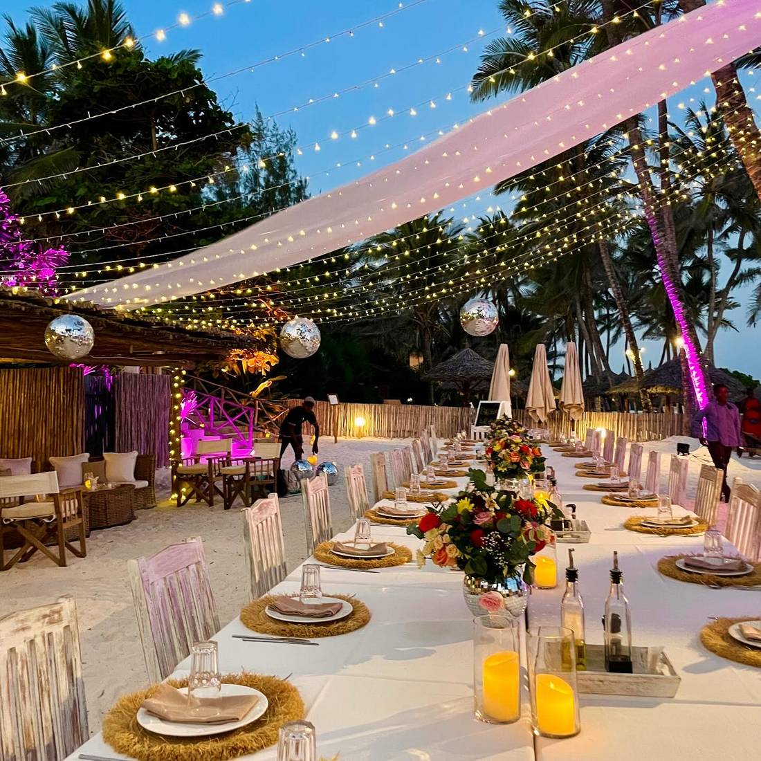 A long banquet table set under fairy lights on the beach at dusk for a Tamu event